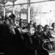 Tom Gerun and members of his orchestra are seated in a roller coaster car at Lakeside Amusement Park in Lakeside (Jefferson County), Colorado. Tom Gerun and two women sit in the front of the car. The musicians are crowed in the rear seats and have their instruments in hand.