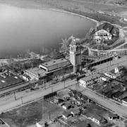 Aerial view of Lakeside Amusement Park in Lakeside (Jefferson County), Colorado. The roller coaster tracks encircle the dance hall and run along the lake (Lake Rhoda). The Star ferris wheel, Casino Tower entrance, a pavilion, and neighborhood houses are shown.