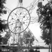 A view of the Staride ferris wheel at Lakeside Amusement Park in Lakeside (Jefferson County), Colorado. A man sits in a ticket booth at the base of the ride. Lake Rhoda is in the distance.