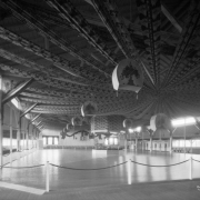 Interior view of the El Patio ballroom at Lakeside Amusement Park in Lakeside (Jefferson County), Colorado. An octagon-shaped bandstand is in the center of the large room with a wood parquet floor. Chinese-style lanterns painted with Art Nouveau floral designs hang from the ceiling along with fabric runners which radiate from two central points. Folding chairs line the room.