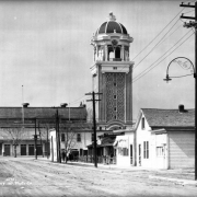 View of the Casino Tower at Lakeside Amusement Park in Lakeside (Jefferson County), Colorado. The contours of This elaborate Italianate domed structure are covered with light bulbs that are strung in geometric patterns. A sign above the door reads: "Lakeside Park" and is covered in light bulbs. Homes and commercial buildings block the view of the entrance. A sign outside a store reads: "Groceries," and a Bell Telephone sign hangs on a utility pole. A man in the street tips his hat to a woman and child.
