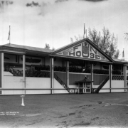 Exterior view of the Fun House at Lakeside Amusement Park, Lakeside (Jefferson County), Colorado. The building has French colonial features including paired exterior staircases, a pavilion roof and thin wooden colonettes. Art Deco designs decorate the columns and the sign reading "Fun House." A face mask and smiling clock are located on the gable. A clown head is attached to the porch, and a monkey figure sits on top of the ticket booth.