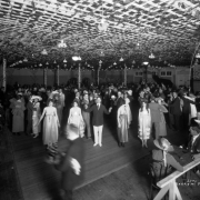 Dancers crowd the floor of the El Patio Ballroom at Lakeside Amusement Park in Lakeside (Jefferson County), Colorado. A man and four women in satin dresses are lined up in one corner of the room while couples occupy the rest of the floor and the tables surrounding dance area. The hall is decorated with an ivy covered trellis and tulip light fixtures.