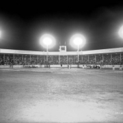 Spectators fill the grandstand of the softball park at Lakeside Amusement Park in Lakeside (Jefferson County), Colorado. Players sit on benches under the floodlights. Men in suits are near first base. A press box sits on top of the grandstand.