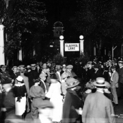 Nighttime view of men and women who crowd around the edge of a sidewalk referred to as "Flapper Lane" at Lakeside Amusement Park in Lakeside (Jefferson County), Colorado. Most of the women have short bobbed haircuts, and the men wear caps and straw hats. Two girls walk arm-in-arm, one wears knickers. A policeman stands next to a column. A tower in the distance is dimly illuminated by lightbulbs.