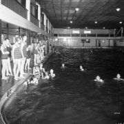 Boys and girls stand at the edge of and swim in the swimming pool at Lakeside Amusement Park in Lakeside (Jefferson County), Colorado. The boys and girls both have full bathing suits, and many of the girls wear bathing caps.