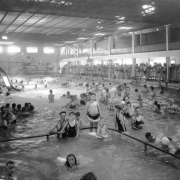 Men, women and children swim in the indoor pool at Lakeside Amusement Park in Lakeside (Jefferson County), Colorado. A girl prepares to go down a slide, and boys and men line up on a platform to use the diving board. Spectators lean against a fence.