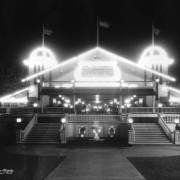 Nighttime view of the El Patio ballroom at Lakeside Amusement Park in Lakeside (Jefferson County), Colorado. Lights illuminate a pair of towers, eaves and the gable end of the building where a sign reads: "El Patio Dancing." Staircases lead to the main dance floor. The interior is decorated with wrought iron grating and Norman style light fixtures. A fountain is in front of the building.