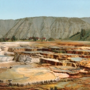 View of Hymen Terrace at Mammoth Hot Springs, Yellowstone National Park, Wyoming. Shows thermal pools and terraces, the Liberty Cap, an extinct hot spring cone, and two-story buildings with red roofs, probably Fort Yellowstone.