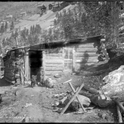 Shows a miner sitting in doorway of his log cabin (lean-to) built into mountainside with glass window frame; includes two dogs, various tools around entrance, wood chopping area, and other mine buildings and structures in background.