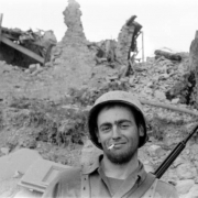John W. Haines, III, of Company I, 87th Mountain Infantry Regiment, Tenth Mountain Division, faces the camera and smiles with a cigarette in his mouth and a rifle on his back.  Behind him are the ruins and rubble of Pietra Colora. Haines was later killed in action.