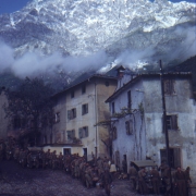 Shows a long line of more than thirty Tenth Mountain Division soldiers waiting for food. The chow-line snakes up a steep street in an Italian town; a few men are eating around a covered jeep.  Above the town is a rugged mountain covered with snow; clouds hover over the roofs.