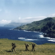 A trio of soldiers from the 87th Mountain Infantry Regiment on patrol loaded with full rucksacks and rifles crosses the muskeg. The sea is beyond them. View is toward the north from near Cape St. Stephen.