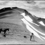 View of summit at Treasure Mountain, Gunnison County, Colorado, with L.C. McClure behind his camera & tripod; horse stands beside unpacked saddle bags; additional camera case, equipment, and probably crate of glass plates are near McClure; clouds top the peaks.