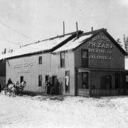 A wagon drawn by a team of horses is parked next to a brewery in the town of Creede, Colorado, in Mineral County. The driver sits in the cart and a man stands next to the wagon. The building is a two-story, wood- frame structure with gables, shingles, chimneys, rectangular windows, and a covered porch with spindles. Snow covers the ground and the hills in the background. A "Beer Depot of P.H. Zang Brewing Co. Jno. Knodel, Agt." shows.