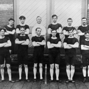 Colorado City Hose Team pose in front of the diagonal batten door outside the brick fire station, South Twenty-sixth and Cucharas streets, El Paso County, Colorado. The men stand wearing dark underwear shirts and shorts with arms crossed and their brass hose nozzle proudly on display in front of them.