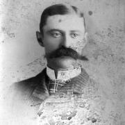 Head and shoulders studio portrait of Jim Boughton with cropped hair parted on the side and a bushy, handlebar moustache, Aspen, Colorado; dressed in a buttoned, tweed suit jacket with a high-collared shirt, and a print tie.