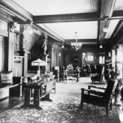 Interior view of the long reception hall in the Frederick Warshauer residence in Antonito, Conejos County, Colorado; includes Arts and Crafts furniture, chairs, tables, benches with leather pillows, lamps, hanging lanterns, oriental rugs, bronze sculptures, and potted palm plants; beamed ceiling with a stenciled border and stairway leading to the second floor.