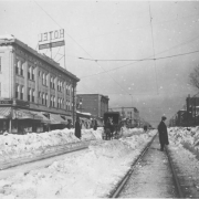 View of Broadway at Ellsworth Avenue after a heavy snowfall in Denver, Colorado. Pedestrians and a horse-drawn buggy are on the street. Signs read: "Hotel" and "No[?]th Drug Co".