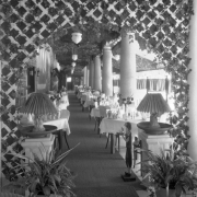 View of a restaurant at Lakeside Amusement Park in Lakeside (Jefferson County), Colorado; shows dining tables with tea service, flower vases, lattices, columns, ivy, potted plants, and ornate lamps. A wooden silhouette women in flapper costume holds an ashtray.