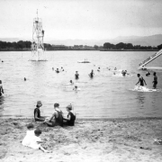 Teenagers and girls play in the sand by Lake Rhoda at Lakeside Amusement Park in Lakeside (Jefferson County), Colorado; a lifeguard on a tower watches swimmers.