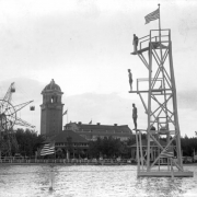Men stand on a diving tower in Lake Rhoda at Lakeside Amusement Park in Lakeside (Jefferson County), Colorado; United States flags, the pavilion, and a ferris wheel are in the background.