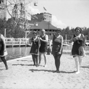 Women and a man walk in sand by Lake Rhoda at Lakeside Amusement Park in Lakeside (Jefferson County), Colorado; they wear swimming suits. One woman plays a ukelele.