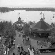 View of Lakeside Amusement Park in Lakeside (Jefferson County), Colorado; people walk by a pavilion and the boat house. Lake Rhoda and Lakewood are in the background.
