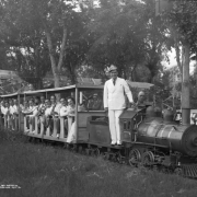 Jimmy Garrigan stands on a miniature locomotive at Lakeside Amusement Park in Lakeside (Jefferson County), Colorado; men and women with a guitar, saxophone, trombones and drums ride passenger cars.