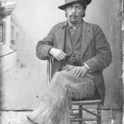 Portrait of August Von Rissling seated in a wooden chair in Leadville, Colorado. Possibly taken outside on dirt with a drop painted with a balustrade. Von Rissling wears chaps with fringe, a sack jacket and vest and a hat tilted on his head. His hair hangs out from under the brim of his hat and he wears a mustache.