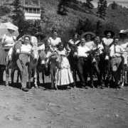 Young men, women, and burros pose in a clearing at the resort in Eldorado Springs, Colorado. Each of the eight burros has a woman on its back, and the men hold the necks of, and stand between, the burros.