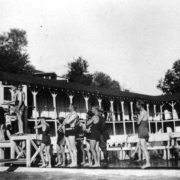 Men and women stand in line at the diving board at the swimming pool in Eldorado Springs, Colorado. Viewing areas and changing rooms surround the pool.