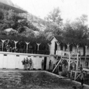 Swimmers and onlookers are at the pool in Eldorado Springs, Colorado. One man is on the lower of two diving boards, and three men stand  near the changing rooms.
