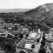 View of the resort at Eldorado Springs, Colorado, includes swimming pools, access to South Boulder Creek, Eldorado Hotel, and a dance pavilion (with barrel roof). Cars are parked all along the dirt road. Crowd  of people are in and around the pool.