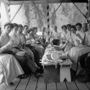 Young men and women sit around a make-shift table of wooden benches that have been pushed together. They have just finished a meal and their empty plates litter the top of the bench. Many are holding up their glasses in the gesture of a toast.