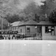 Four women and a young girl are poised to dive into the swimming pool at the resort in Eldorado Springs, Colorado. A fifth woman stands on the running board of a bus behind the divers. Signs on the bus read: "Inter-urban motors - the Boulder Line." A man leans over the railing in the viewing area, and women and children are behind him. A clock on the wall of the resort office building reads 6:10.