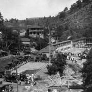 Crowd of people gather around the swimming pool at the resort in Eldorado Springs, Colorado. South Boulder Creek is in the foreground and has a bridge over it. Other people walk from their cars towards the pool. A  building with the sign "Grand View"  is in the distance.