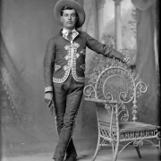 Studio portrait of a man. He holds a pistol and wears a jacket and pants decorated with embroidery and a wide-brimmed hat.