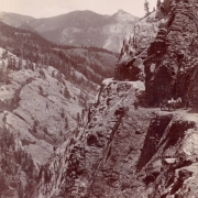 A driver in a horse-drawn buggy and a man on horseback stop on the Otto Mears toll road on a high cliff probably Ouray County, Colorado. Shows talus and rock formations, the narrow road is buttressed with logs. A passenger sits by the road.