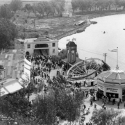 People walk by a stage, clock tower, mechanical ride, Lake Rhoda, and a concession stand with sign: "Refreshment Parlor" at Lakeside Amusement Park in Lakeside, Colorado.