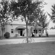 Men and women stand and walk near a church probably in Denver, Colorado. The one-story brick building has a curvilinear gable, modillions, and tile awnings. A bicycle is nearby.