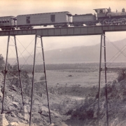 View of a Colorado Midland locomotive, baggage and mail car, and excursion car high on a railroad trestle in a gorge near Buena Vista, Chaffee County, Colorado. Shows employees of the rail company on the train. A wood frame building is in the valley