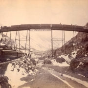 View of a Colorado Union Pacific, Denver & Gulf Railroad freight train on the narrow gauge High Bridge trestle near "The Loop," a route over Clear Creek near Georgetown (Clear Creek County), Colorado, men stand on top of the freight cars. A passenger train is on a rock rail embankment below the bridge, passengers and rail workers stand on rocks near the creek; snow-covered mountains are in the distance.