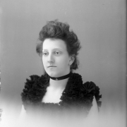 Studio portrait of a woman. She wears a sleeveless dress and a choker.