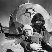 A woman holds a child bundled in a coat, hat, and mittens in Drop  City, Colorado, in Las Animas County.  They stand outside of a geometrically-shaped dwelling (geodome) with pentagon-shaped windows.  A coarse, wood ladder leans against a conical-shaped structure in the background.