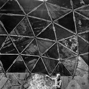 A man stands in the interior of a partially-finished geodesic dome in Drop City, Colorado, in Las Animas County.  Light streams through the geometrically-shaped window at the top of the structure.  The man holds a circular painting with his right hand and stands amidst other pieces of art on the floor.