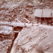 Men pose near a toll gate on the Otto Mears toll road in Ouray County, Colorado. A horse-drawn buggy, wood frame building, and a man on horseback are nearby. Bear Creek Falls is under the bridge.