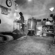 Interior of a geodome in the community of Drop City, Colorado, in  Las Animas County; shows bed covered with blankets on floor, kitchen area including refrigerator and sink, a clock on wall set to 1:10, and light through triangular-shaped window on ceiling.