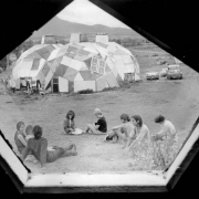 View taken from inside a geodesic dome looking outside at people sitting on the ground in Drop City, Colorado, in Las Animas County.  Three geodesic dwellings are in the background and have cars parked next to them.