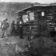 Five miners stand outside of a log cabin in Dunnville, Colorado.  Tents are in the background and debris and garbage litter the ground. Posters are attached to the side of the cabin.